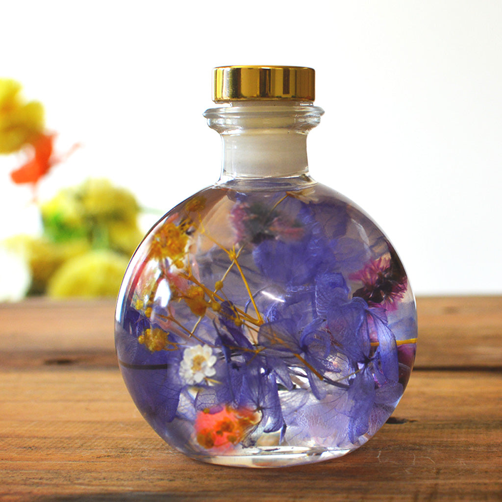 Plum Streak Tabletop - A glass bottle with a cork containing preserved plants with a mix of purple, pink, and yellow flowers, displayed on a wooden surface.